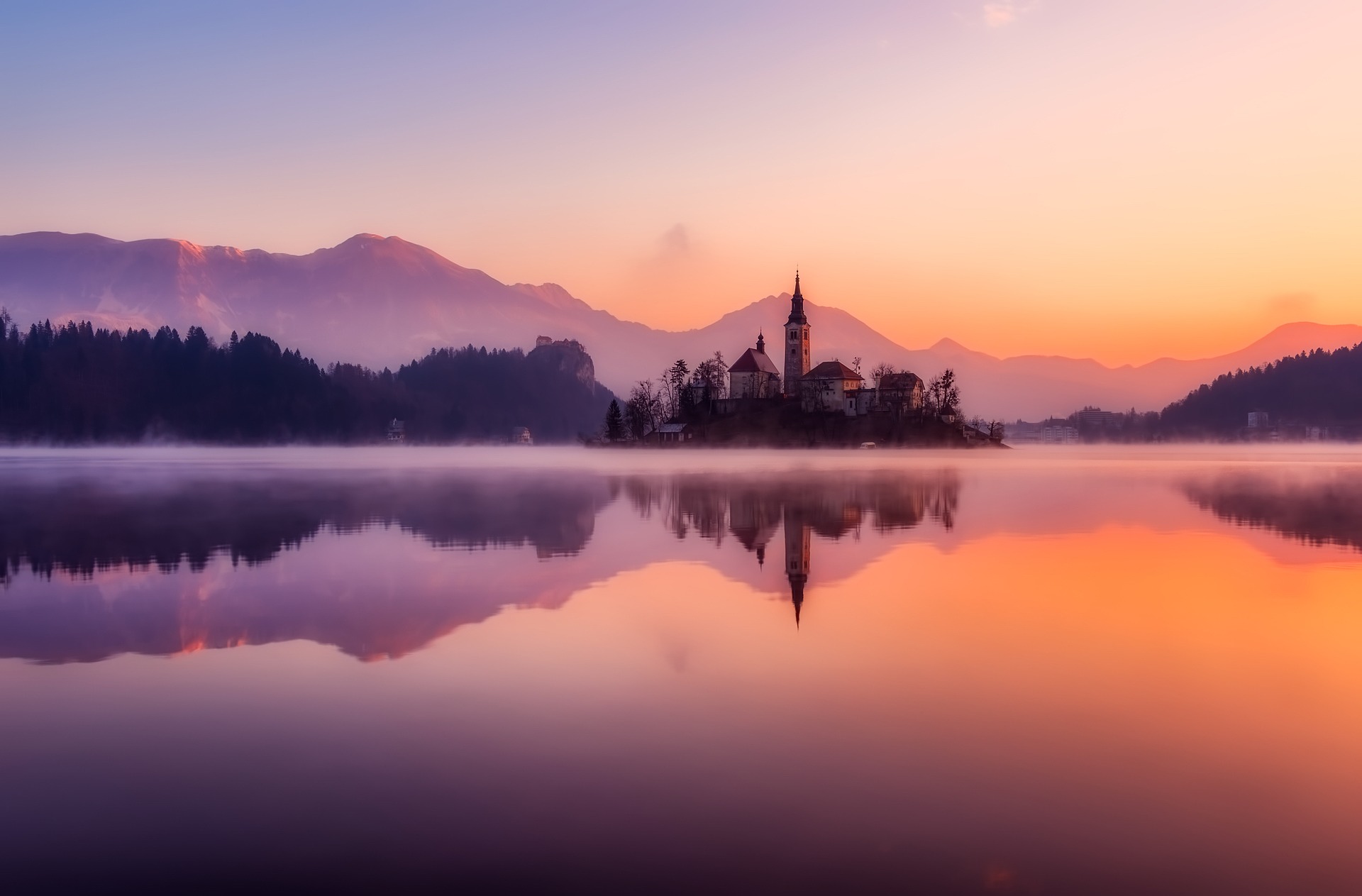 Scenic lake at dawn with misty water, a small island with a church tower, and mountains in the background under a pastel sky