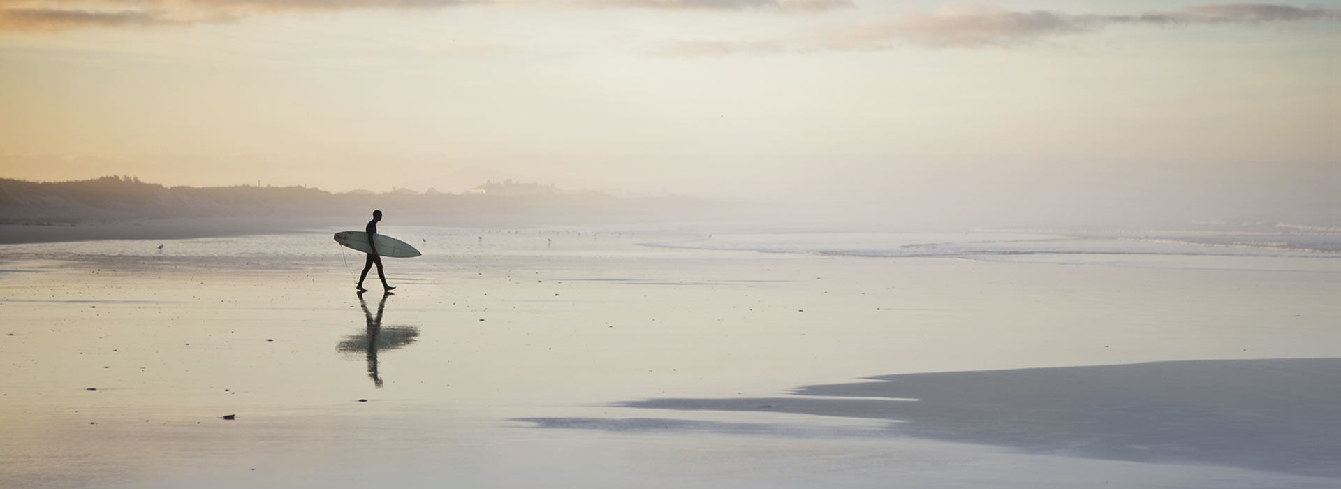 Silhouette of a person carrying a surfboard walking along a calm beach at sunset, soft pastel tones