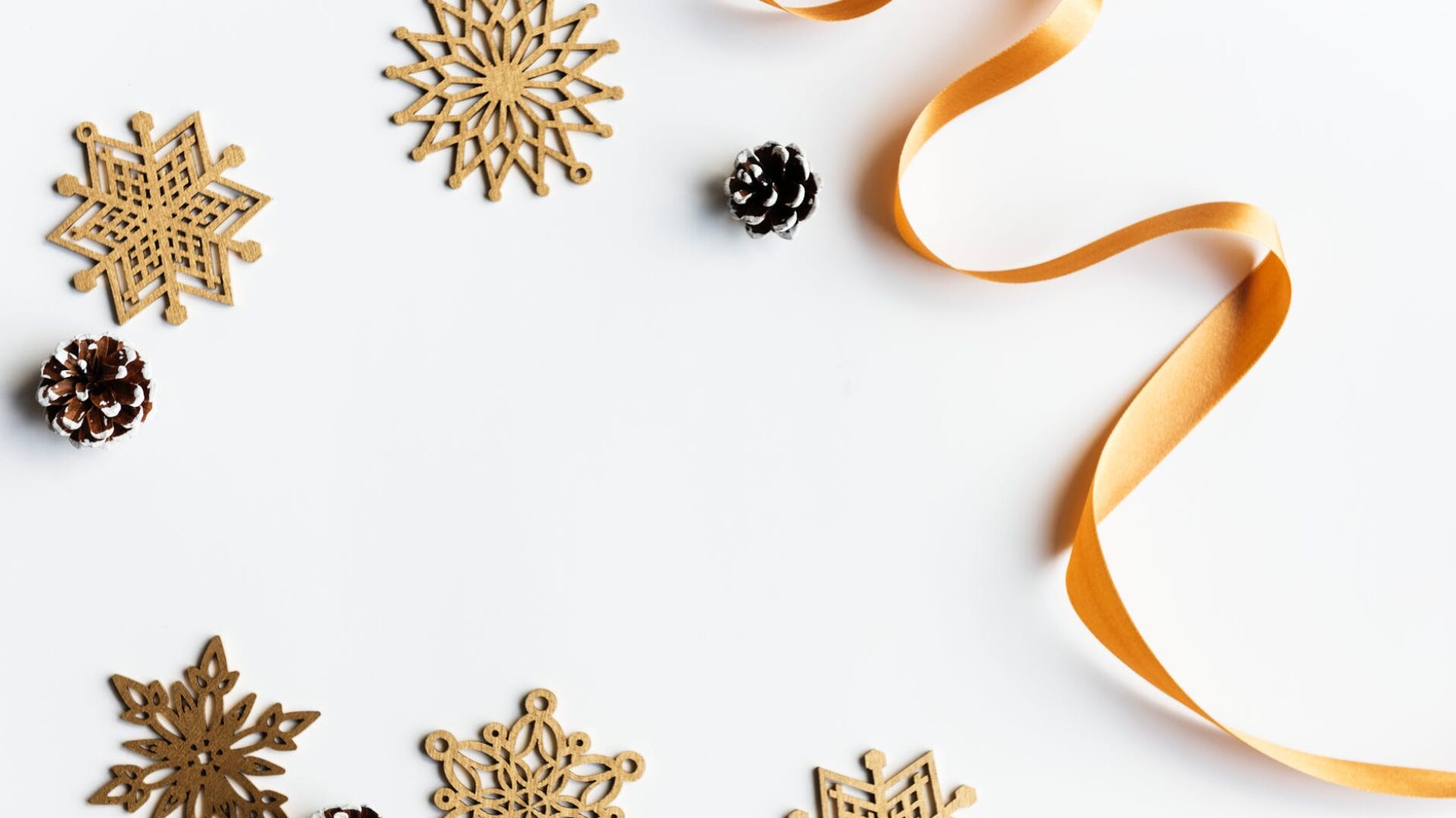 Flat lay of decorative wooden snowflakes, small pinecones, and a curled gold ribbon on a white surface